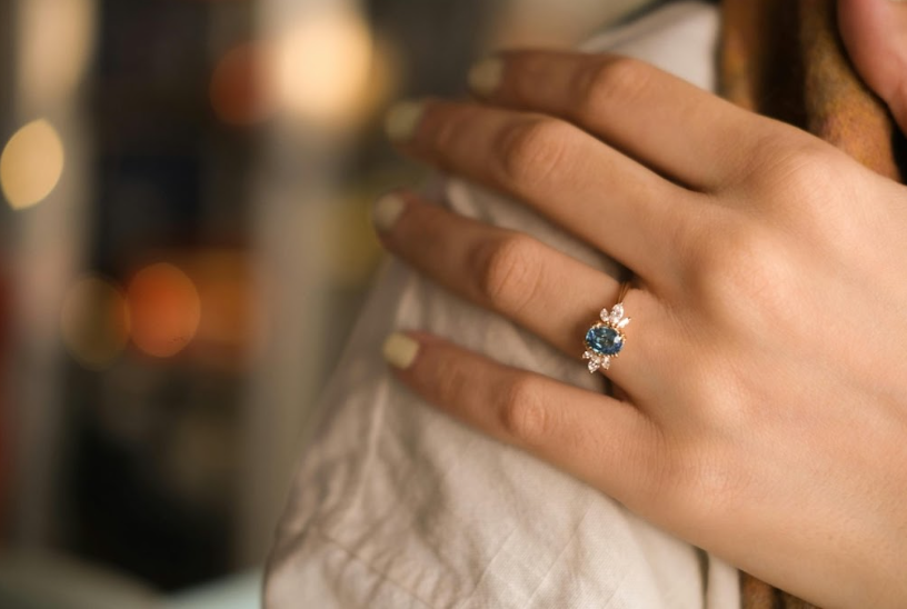 A woman's hand adorned with a yellow gold ring featuring a blue gemstone surrounded by small diamonds, placed on her shoulder against a blurred background.