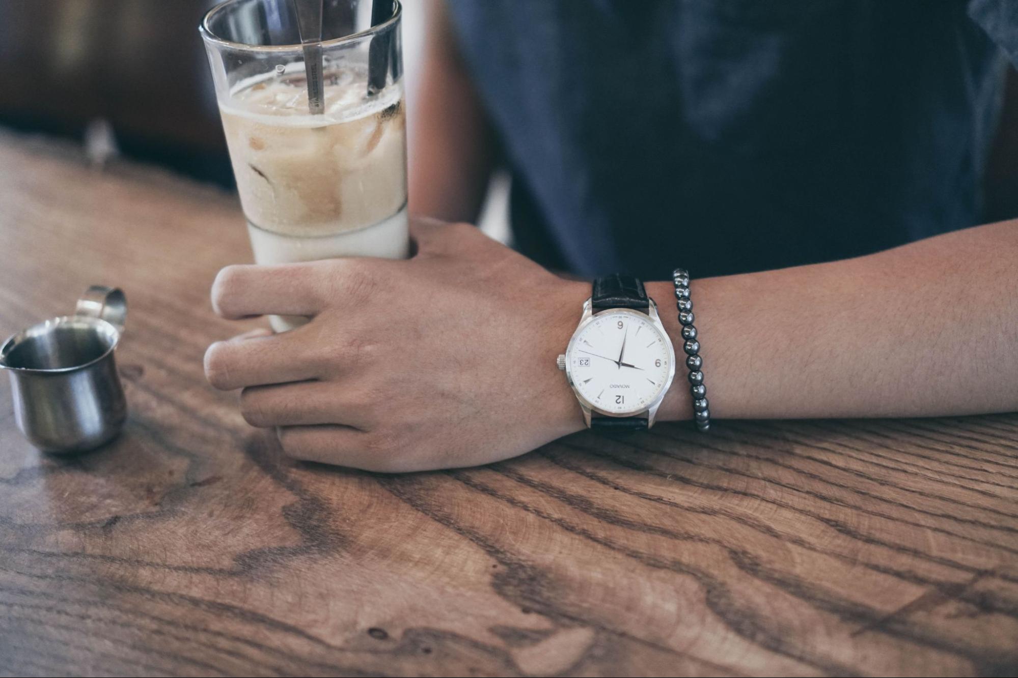 A close-up of a casually dressed man drinking iced coffee while wearing a vintage watch.