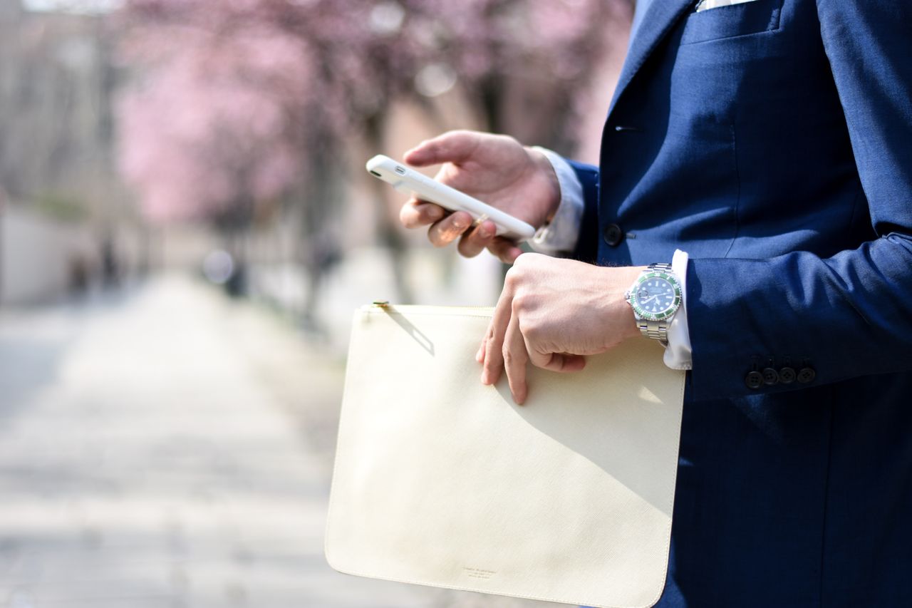 A well-dressed man wearing a luxury watch holding a zipper pouch while checking his phone.