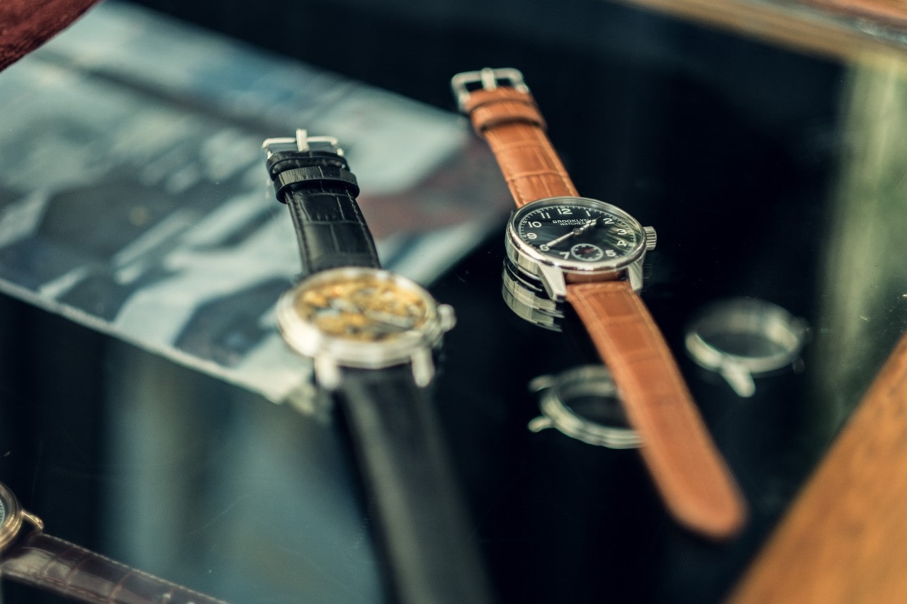 Two luxury watches displayed side-by-side on a glass-top table, a third watch almost out of view.