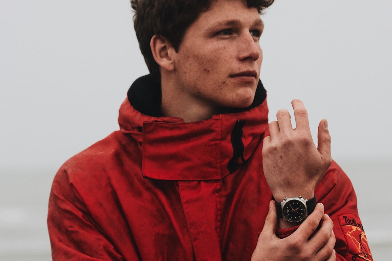 A young man in a red raincoat wearing a black and silver luxury watch.