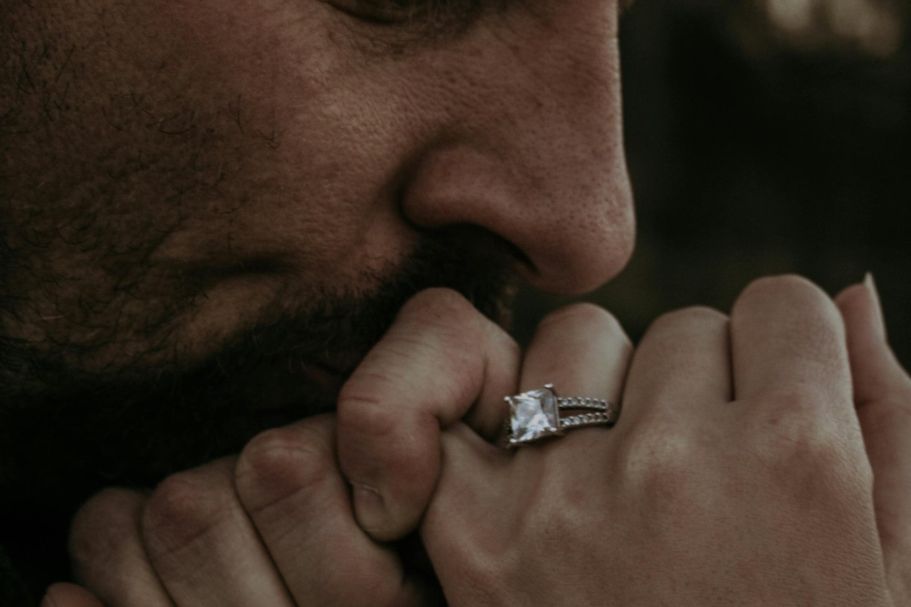 A close-up of a man kissing a woman’s hand, her hand adorned with a princess cut diamond engagement ring.