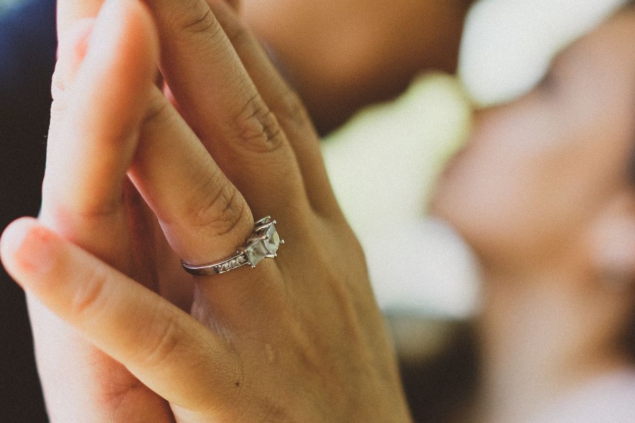 Close up image of a couple’s hands pressed together, the woman wearing a three stone engagement ring, their faces blurred in the background