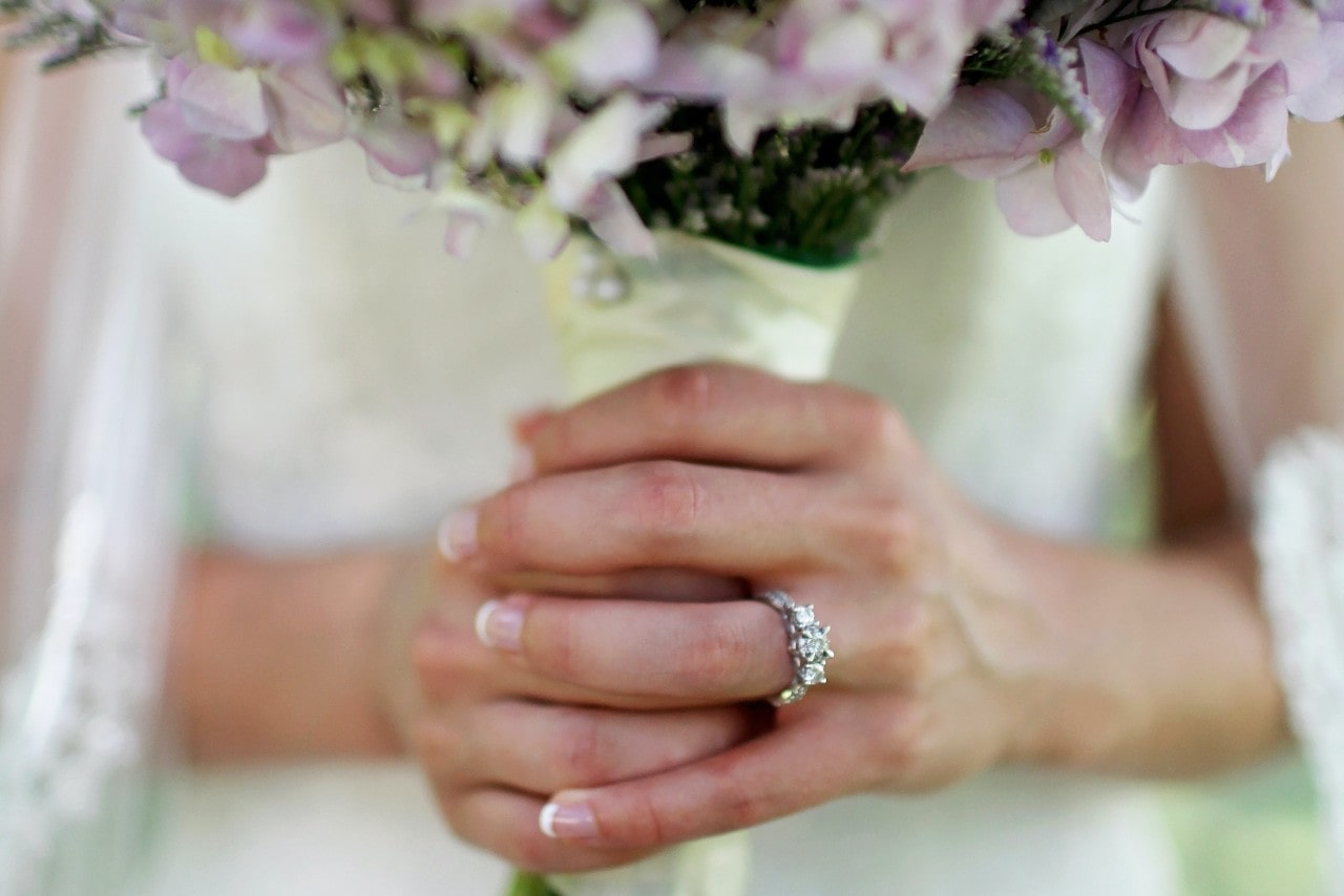 Close up image of a bride’s hands holding a bouquet of flowers and wearing a silver, three stone engagement ring