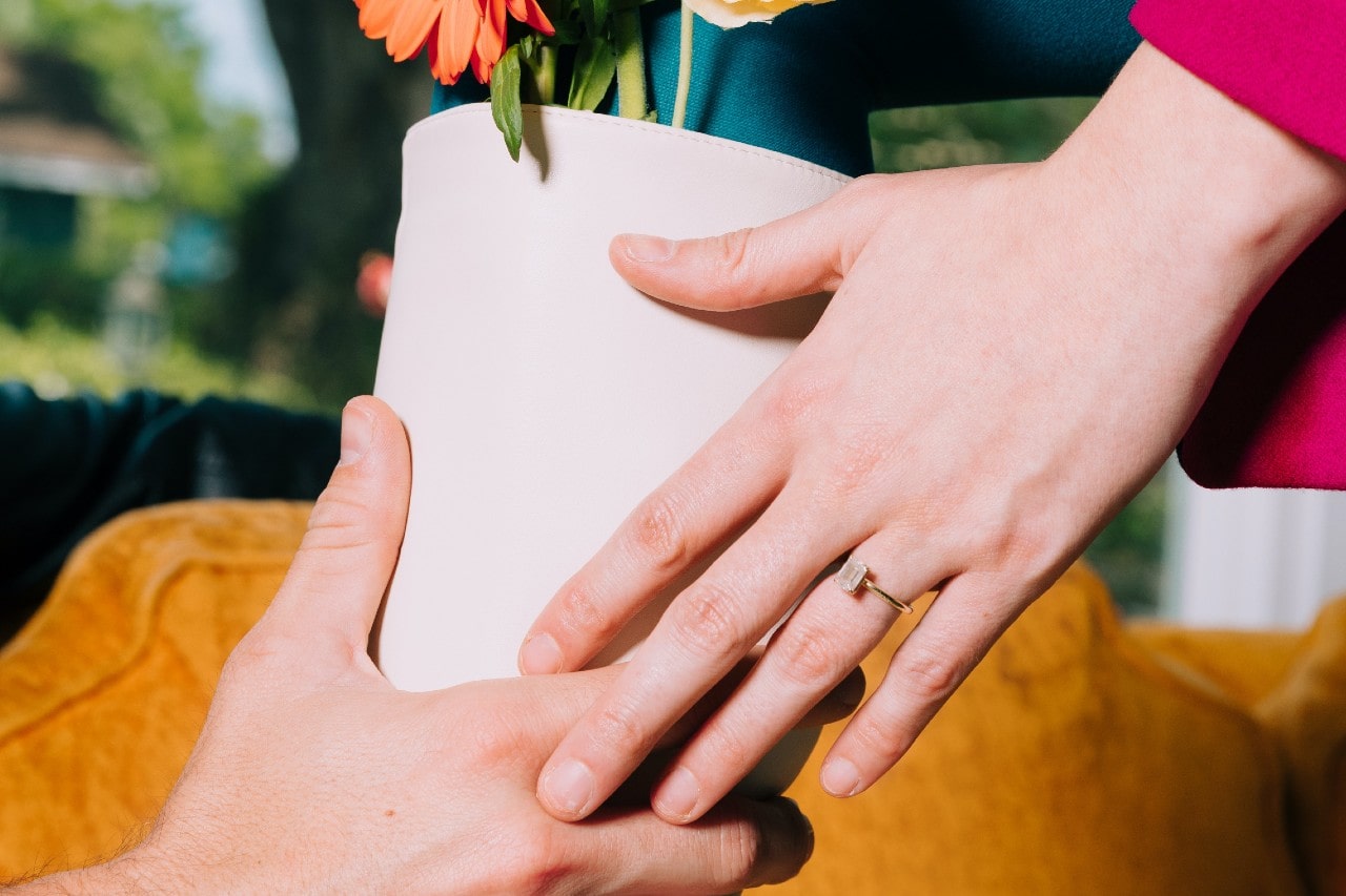 Two people passing a white flower pot— the woman wearing a dainty engagement ring.