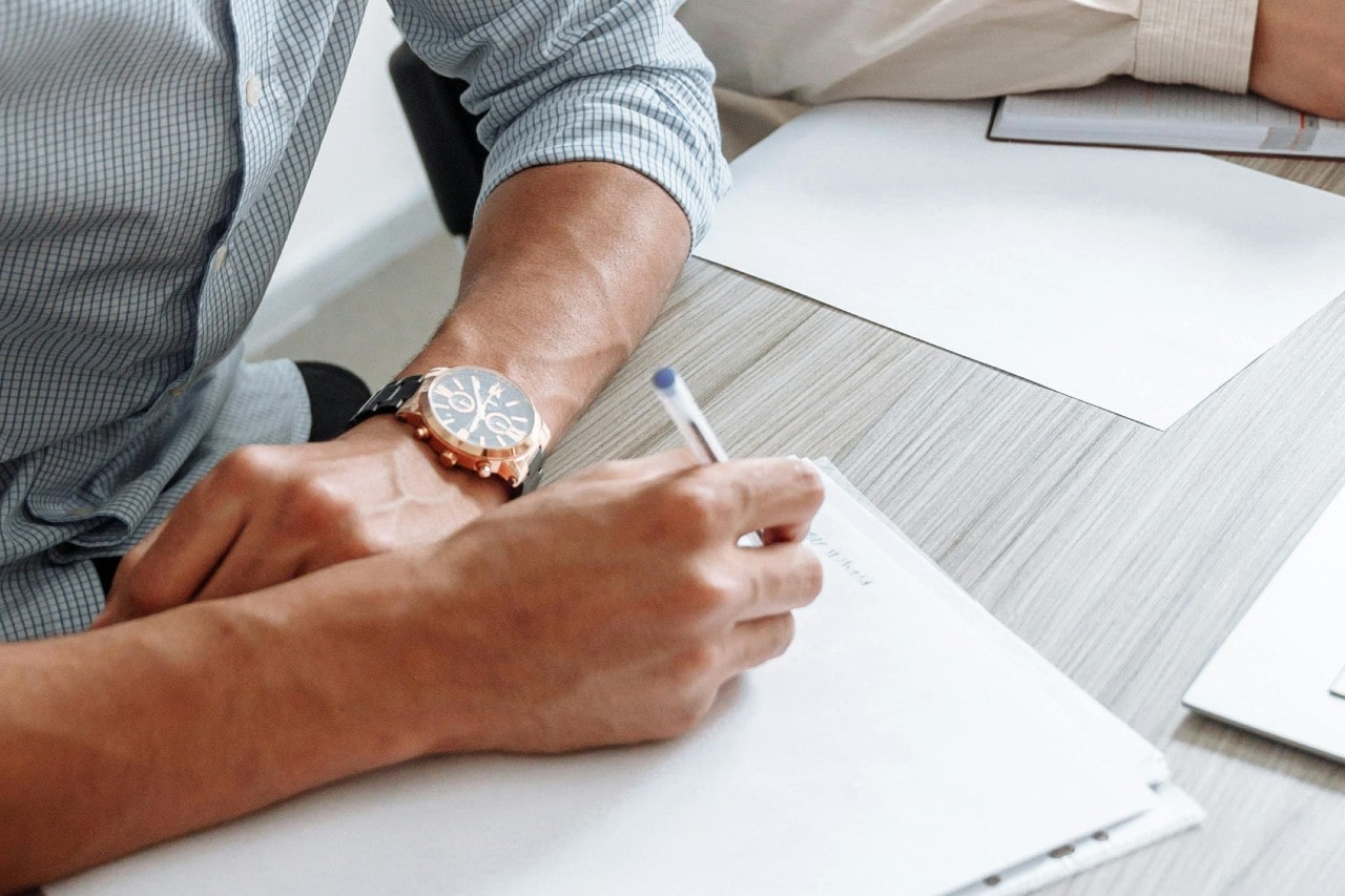 A close-up of a man writing a letter, a gold watch on his left wrist.