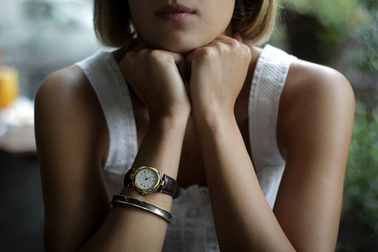 A close-up of a casually dressed woman, her right wrist adorned with a luxury watch and a simple bracelet.