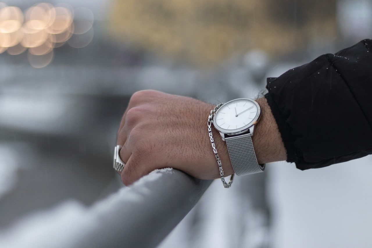 A close-up of a man’s hand on a rail, adorned with silver-toned jewelry and a luxury watch.