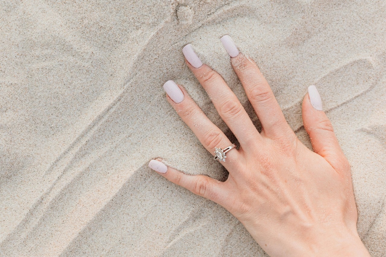 A woman’s hand in the sand adorned with a marquise cut engagement ring and a halo setting.