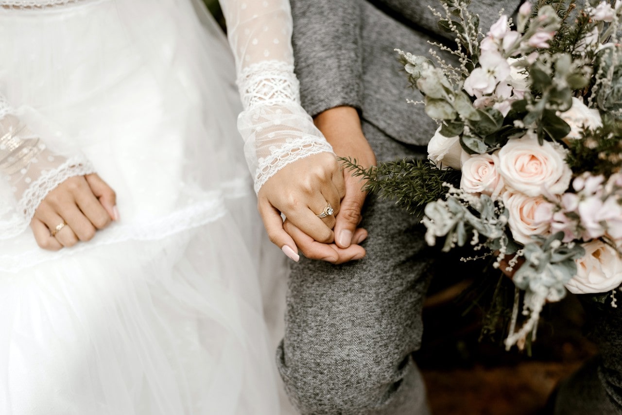  close-up of a newlywed couple holding hands, a two-tone engagement ring visible on the bride’s finger.